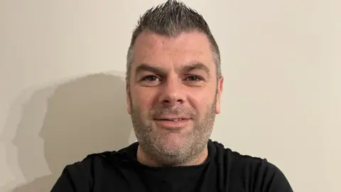 Kevin McMullan A man in a black t-shirt in front of a plain background smiles at the camera