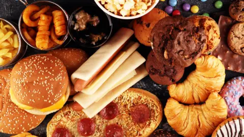 Getty Images An overhead view of food which can be described as ultra-processed, including chocolate muffins, hot dogs, supermarket pizza, crisps, popcorn, doughnuts, burger buns and sweets