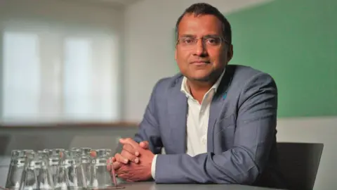 Dr Kremlin Wickramasinghe from the World Health Organization, who has short dark hair and wire-rimmed glasses, sits at a table with his hands clasped together. Some upturned glasses are in front of him on the table and he is wearing a white shirt and suit jacket.
