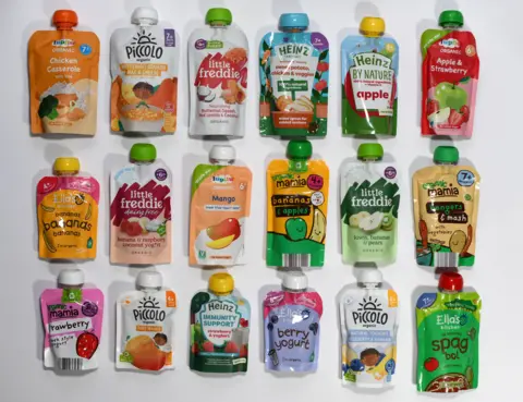 18 baby food pouches lined up in three rows, against a white background. There is a selection of savoury, fruit and yoghurt.