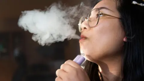 Getty Images A young person exhaling smoke from a vape, which she is holding in her hand. 