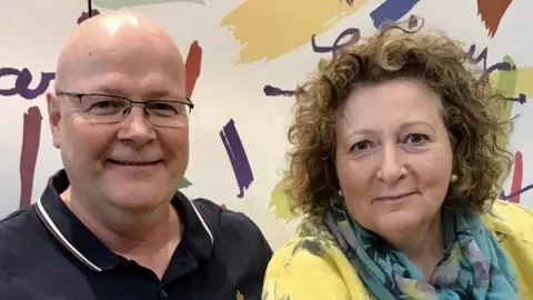 Family handout Stephen and Carol Baxter, a husband and wife who are looking directly at the camera and smiling. Stephen Baxter is wearing black rimmed rectangular glasses and is wearing a blue polo shirt. Carol Baxter is wearing a yellow floral top with a blue floral scarf. She is wearing pearl earrings and has brown curly hair.