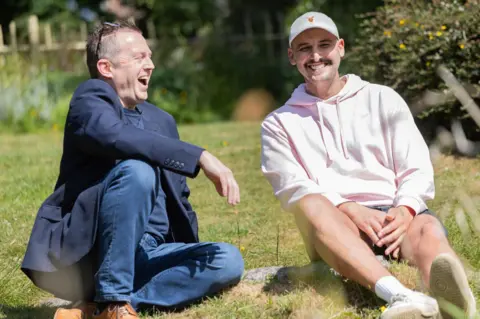 Caters Photographic Two men sitting on grass in a park. On the left, a 51-year-old man wearing a blue blazer and blue jeans smiles broadly as he looks at a 31-year-old man who is smiling and wearing a white cap, a light-pink hoodie and shorts.