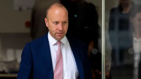 PA Media Matt Hancock leaving the Covid inquiry on 22 May. He has a shaved head and is wearing a dark blue suit, a white shirt and a pink tie. 