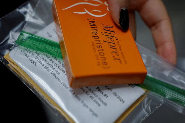 A hand holds a small orange box of Mifepristone along with a plastic bag containing typed directions.