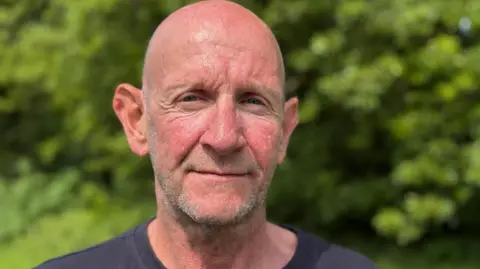 BBC Lee is stood looking at the camera, wearing a dark tshirt. He is shaven headed with short, stubbled facial hair. He is outdoors on a sunny day, with a green hedge blurred in the background.