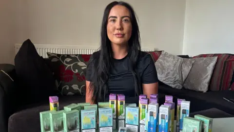 Cara Hallinan Cara is a 25-year-old woman with long dark hair, she is wearing a black T-shirt and she is sitting on a sofa. On a table in front of her is an array of multi-coloured disposable vape boxes.