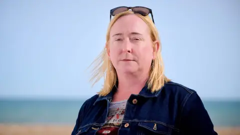 BBC A woman with blond shoulder-length hair, standing on a pebble beach with the sea in the background. She is wearing a blue denim jacket. The sky is blue and cloudless.
