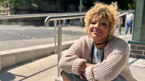 BBC / Hazel Shearing Vika Zak sits on some steps outside a building in Nottingham. The sun is shining and she's wearing a neutral cardigan with a blue t-shirt underneath. She has a nose ring, a lip ring and a wears a bracelet on her right wrist.