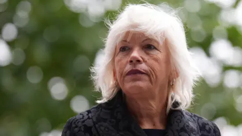 PA Media Melanie Leahy has white shoulder-length hair, and is wearing a dark jacket with floral pattern. She has small silver hoop earrings and wears dark red lipstick. She stands in front of a blurred background of trees