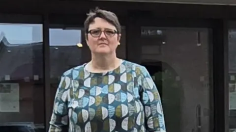 A woman with short dark hair and dark glasses, stood in front of a glass building. She is looking into camera and wearing a green, blue and white top.