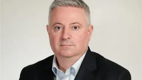 ESSEX PARTNERSHIP UNIVERSITY NHS FOUNDATION TRUST Current ceo of EPUT Paul Scott. He is looking directly at the camera and you can see his head and shoulders. He is wearing a black suit jacket and checked white and pale blue shirt. He has grey hair.