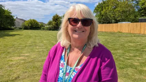 Jan is smiling at the camera as she stands in the large lawned grounds of Brynawel House on a sunny day. She is wearing prescription sunglasses and a floral top and pink cardigan.