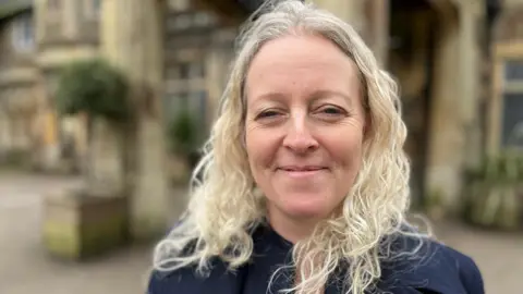 Julie has long, curly blonde hair and is smiling at the camera. In the background is the venue where the Everywoman festival is held.