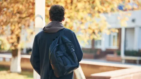 Getty Images A student walks away from the camera. You can't see his face, but he has brown hair and wears a hoodie with a grey jacket and a black backpack.