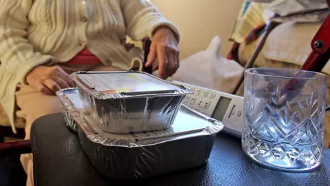 Getty Images Two foil food containers stacked, and empty glass and a landline wireless phone sit on a black leather-like surface. Behind it is an elderly person wearing a beige cable knit cardigan with buttons. Face is not visible and clothing does not make it possible to distinguish if they are male or female.