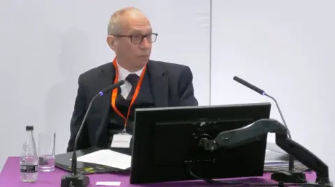 Lampard Inquiry Sir Rob Behrens appearing at the Lampard Inquiry. He sits at a purple-covered desk with two microphones pointing towards him and a computer monitor in front of him. He has a bottle of water and a glass to his right hand side. He is wearing a dark suit with dark waistcoat, dark tie and white shirt. he has dark-rimmed glasses.