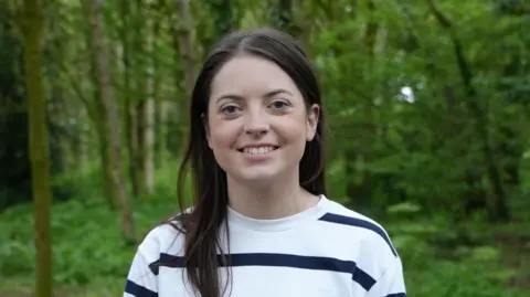 BBC Kathryn is stood in a wooded area, she is wearing a white striped jumper and has long, dark hair.