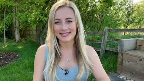 BBC Eve Morgan, blonde and smiling at the cameras earring a grey vest and small pendant necklace. She is sitting in a garden area with grass and trees behind her