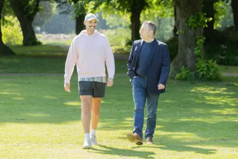 Caters Photographic Two men walking on grass in a park on a sunny day. On the left is a tall man wearing a light-pink hoodie, a white baseball cap and shorts. He has a dark moustache and is smiling broadly. On the right, an older man is wearing a blue blazer and blue jeans and is looking at the other man.