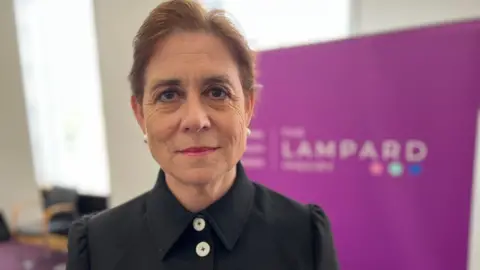 STUART WOODWARD/BBC Baroness Kate Lampard looks straight at the camera. She is standing in front of a purple 'Lampard Inquiry' sign. She is wearing a black blouse with a large collar and large buttons, her hair is short and she is wearing pearl earrings.