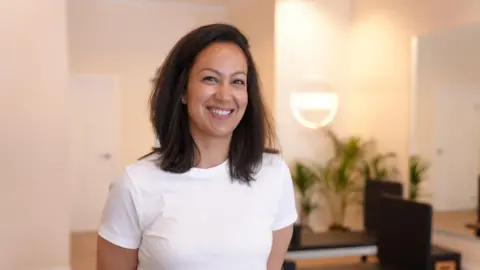 Shakira is in her pilates studio, with reformer machines in the background. She is wearing a white tshirt and has shoulder-length dark hair.