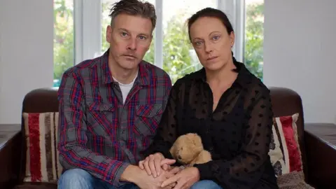 BBC Richard Stanton and Rhiannon Davies sit on a brown sofa with a window behind, holding hands with a teddy bear in Rhiannon's lap