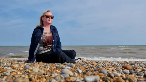 A woman with blond shoulder-length hair, wearing sunglasses, who is sitting on a pebble beach close to the water's edge. The sea is behind her and the sky is blue. She is wearing a blue denim jacket and a top with a big red rose on it.