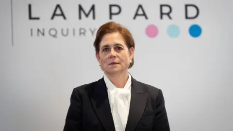 PA Media Baroness Lampard stands in front of a white background with the words Lampard Inquiry written on them. She has dark brown hair with a side parting, large white pearl stud earrings and has dark red lipstick. She is wearing a black blazer jacket and a white blouse.