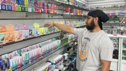 Ruth Clegg Nick is wearing a grey T-shirt and is restocking the shelves in his vape shop. There are rows of brightly-coloured disposable vape boxes alongside him