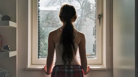 Getty Images Stock image of a girl with long hair tied in a ponytail stands next to a window and looks outside, wearing a vest and checked pyjama bottoms.