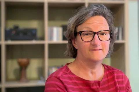 A head and shoulders image of Sarah Hendrickx, who is smiling, wearing glasses and in front of a set of shelves