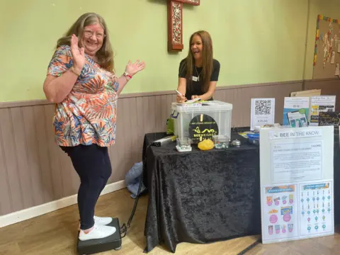 A woman stands on a weighing scale and smiles and waves at the camera. A second woman, Lynda Leadbetter, laughs too as she writes a note down on a piece of paper
