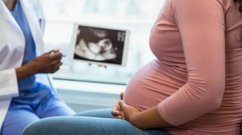 Getty Images A heavily pregnant woman wearing blue jeans and a pink top sits side-on, talking to a healthcare professional who is showing her a baby scan on a tablet.