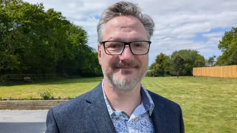 Gareth is smiling at the camera. He is stood with the large lawns of Brynawel House in the background. He is wearing dark rimmed glasses, a navy blazer and a patterned, pale blue shirt.