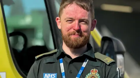 BBC A paramedic wearing a dark green uniform which has an NHS label on. It also has an ambulance badge on it and he is also wearing a blue and white NHS lanyard. The man has auburn hair and and a beard and is smiling at the camera. 