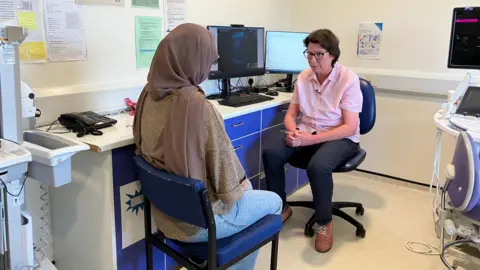 Two women sat in a doctor's office. The doctor is wearing a pink shirt, black trousers and brown shoes and looking at the patient. The patient is wearing a brown hijab, brown top and blue jeans.