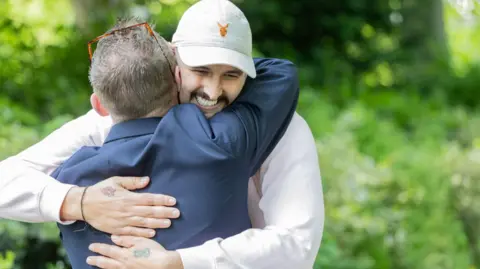 Caters Photographic Two men hug in a park. Facing the camera is a man with a broad smile. He is wearing a light-pink hoodie and a white baseball cap and he has a dark moustache and stubble. His arms arms are wrapped around an older man who has his back to the camera and is wearing a blue blazer and glasses on the top of his head.