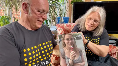 BBC / Hazel Shearing Bob and Maggie Abrahart look at a photo of their daughter, Natasha. They are sitting in their lounge, both on arm chairs.