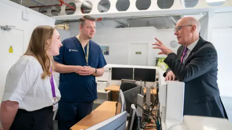 PA Media A woman and a man in medical scrubs speak to John Swinney in a suit leaning over a desk of computers