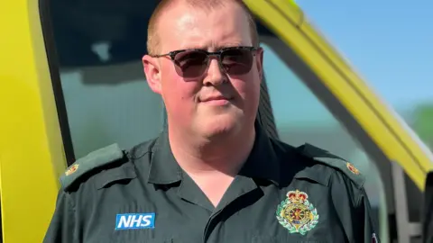 An ambulance worker wearing a dark green uniform looks at the camera. He is wearing glasses and his uniform has an NHS logo and service badge. Behind him the yellow of an ambulance is in view.