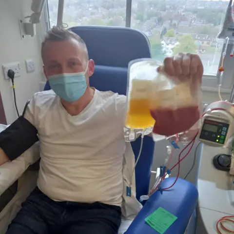 Alastair Hawken A man sitting in a blue hospital chair in front of a window while holding two blood bags, one containing a dark-red substance and the other an amber liquid. He has short light hair and is wearing a blue facemask and a white T-shirt. A black band is strapped around one arm. There is medical equipment next to him.