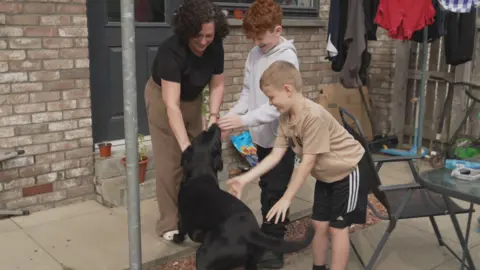 Tracey Meechan, a woman with dark brown curly hair, pets her dog, a black labrador in her back garden, with two of her sons beside her. There is a garden table and chairs beside them and washing is hanging on a whirlygig behind them