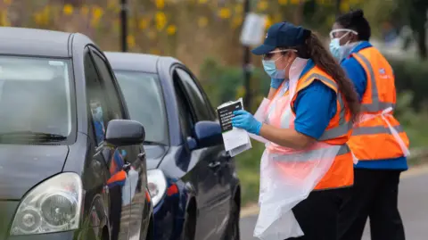 PA Media A drive-in testing site in Southwark, London on 16 September 2020. There are two dark cars queuing to pick up testing kits from staff wearing masks, aprons and gloves. 