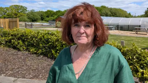 Sue has shoulder length reddish brown hair and is wearing a V-neck green, linen top and silver necklace. She is stood in the grounds of Brynawel House on a sunny day, with the polytunnel in the background where horticultural therapy takes place.
