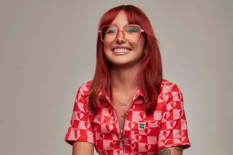 Luke Nugent (Luke Nugent Studio) Ellie Middleton, who has long, red hair and is wearing glasses, is smiling against a plain grey backdrop