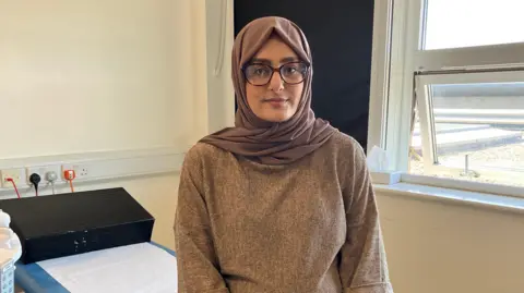 Komal is smiling at the camera, sitting on a bed in a doctor's office. She is wearing black glasses, a brown hijab, brown top and blue jeans.