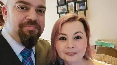 Laura Marston Laura, who has pinkish shoulder-length hair, stands next to her husband, who is wearing a colourful tie and jacket 