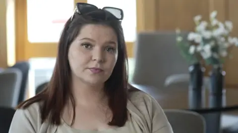 Jamie Niblock/BBC Ellena Baxter a woman with brown hair who is wearing a cream coloured outfit. She is looking towards the camera and is sitting at a table.