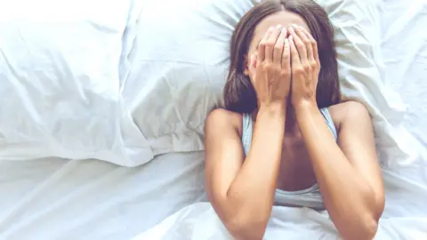 Getty Images A young woman lies in bed trying to get to sleep. She covers her face with her hands. 